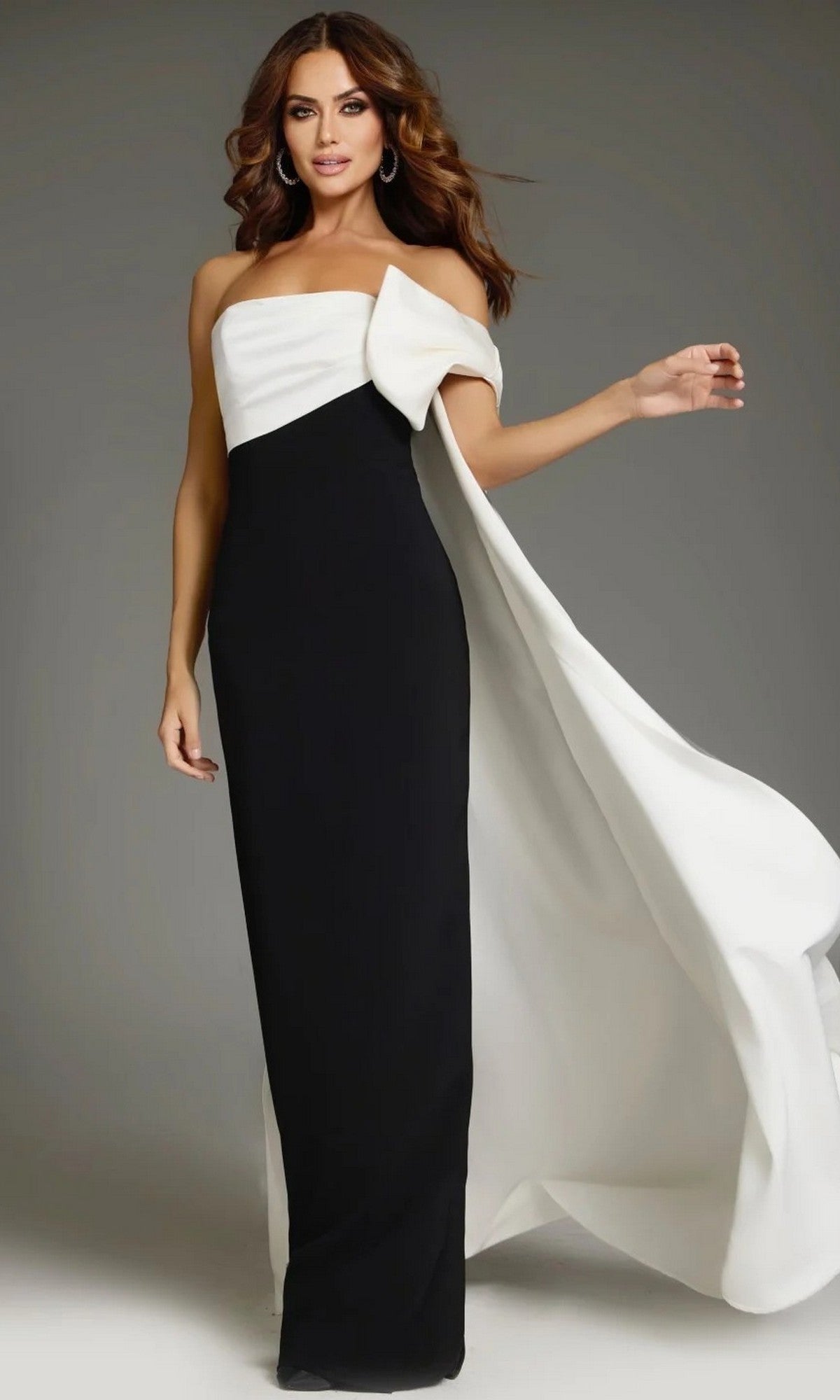 Wearing the Long Formal Dress: Jovani 38864 in black and white, a woman stands against a plain background. This floor-length gown features a strapless design highlighted by a dramatic white drape cascading from one shoulder.